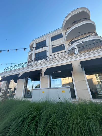 The image shows the facade of a building with a light-colored exterior and curved balconies with ornate black railings. The building has multiple floors with dark-tinted windows and black awnings over the ground-floor windows. A string of lights hangs across the upper portion of the scene. In front of the building, a patch of tall, green grass is visible, and to the right, a grey electrical cabinet with a yellow warning symbol is present. The sky above is clear and blue, with a single, small bird visible in the distance. The building is identified as belonging to "SUNNY ISLAND DEVELOPMENT" and is located in Chernomorets, Bulgaria, likely a commercial or residential establishment.