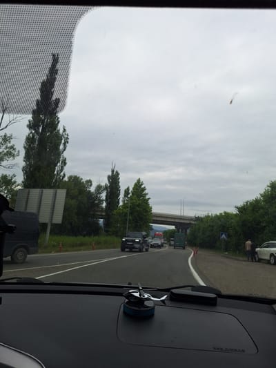 The image shows a view from inside a vehicle, looking out through the windshield. The scene is a road with several cars and trucks, suggesting traffic. Tall trees line the sides of the road, and a bridge is visible in the distance. The sky is overcast. On the dashboard of the car, a small decorative object resembling an airplane propeller and a black case are visible. A sign with the text "SRS AIRBAG" is also visible on the dashboard. The image appears to have been taken on a highway or major road outside of a city. No specific landmarks or indications that this is in Samtredia, Georgia are present in the image.