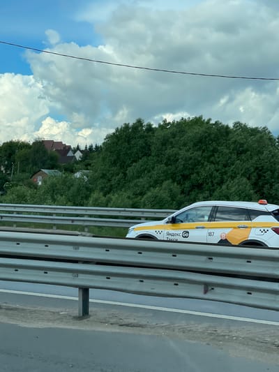 A Yandex Taxi is on the roadside barrier in Bykovka, Russia. The taxi is white with yellow striping and Yandex Taxi markings. A dense green forest is in the background, along with houses with red roofs. A cloudy sky with patches of blue is visible above. A roadside barrier runs horizontally across the lower center of the image. A power line is visible in the sky to the left of the image.