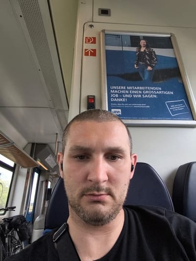 This is a selfie taken inside a train carriage. The primary subject is a man with short hair and stubble, looking directly at the camera. He is wearing a black t-shirt and has wireless earbuds in both ears. Behind him, to his left, a bicycle with a black bag strapped to the rear rack is visible, suggesting he may be traveling with it.

The setting is a train interior with rows of seats. The seats appear to be dark blue or black. Overhead, there is a textured white panel with a perforated section, likely for ventilation or lighting. A silver-colored luggage rack runs along the top of the window.

A prominent advertisement is displayed on the wall to the man's right. The advertisement features a smiling woman in the upper half and German text below. The text, partially visible, includes phrases like "UNSERE MITARBEITENDEN MACHEN EINEN GROSSARTIGEN JOB – UND WIR SAGEN: DANKE!" (Our employees do a great job – and we say: Thank you!) and mentions "Usedomer Bäderbahn" and "starkesTeamUBB". There are also safety symbols, including one resembling a fire extinguisher and an upward-pointing arrow, on the wall above the advertisement.

The lighting suggests it is daytime, possibly late morning or early afternoon, as natural light is filtering in through the windows. The overall mood conveyed by the man's direct gaze is neutral or perhaps slightly casual. The location cues from the advertisement place the scene within the Usedomer Bäderbahn network in Germany.