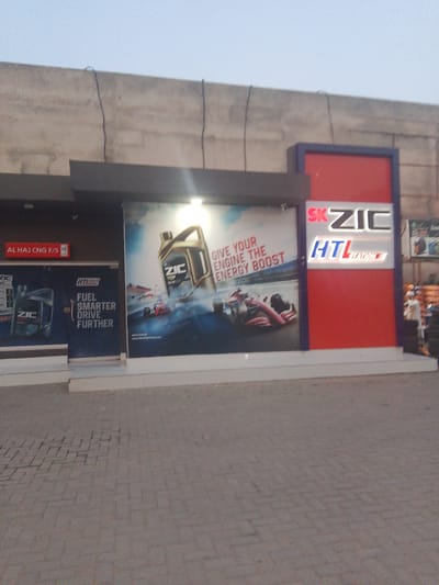 A ZIC HTL Station is visible in Peshawar City Tehsil, Pakistan. The station features a large advertisement with images of ZIC engine oil and race cars, along with the slogan "GIVE YOUR ENGINE THE ENERGY BOOST." A red and blue sign with "SK ZIC HTL STATION" is also prominently displayed. In front of the station, a paved area with brick patterns is visible. To the left, a smaller sign indicates "AL HAJ CNG F/S". A person in a white outfit can be seen in the background on the right, next to stacked tires. The overall scene depicts a commercial establishment for automotive products and services.