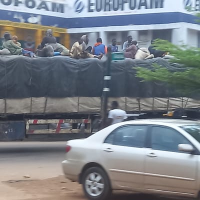 In Sardauna, Nigeria, a white car is parked beside a truck. The truck is crowded with people. The truck appears to be transporting goods. A sign for EUROFOAM is visible. The environment is urban.