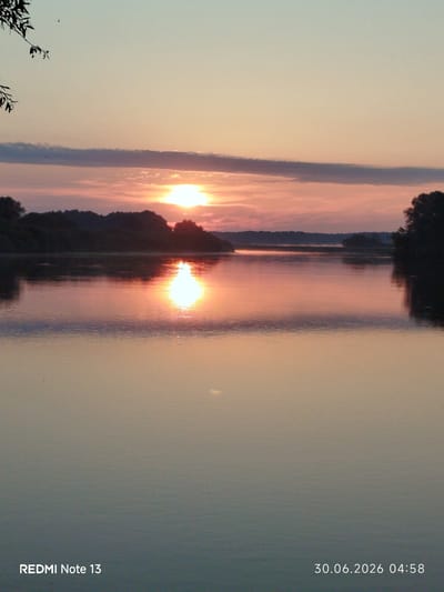 The image shows a serene river landscape at dawn, with the sun just beginning to rise. The sky is a soft gradient of pale blue and pink, with a thin, dark cloud band stretching horizontally across the upper portion. The sun itself is a bright, radiant orb, casting a shimmering golden reflection on the calm surface of the river. Silhouetted trees and a densely wooded shoreline create a dark, undulating horizon line. The water is still, mirroring the colors of the sky and the sun. In the foreground, a branch with a few leaves hangs from the upper left corner, adding a natural frame to the scene. The bottom of the image displays text: "REDMI Note 13" on the left and "30.06.2026 04:58" on the right. This suggests the image was taken with a Redmi Note 13 phone on June 30, 2026, at 4:58 AM. While the exact location within Rahachow, Belarus is not explicitly identified in the image itself, it is understood to be a scenic river view within the city.