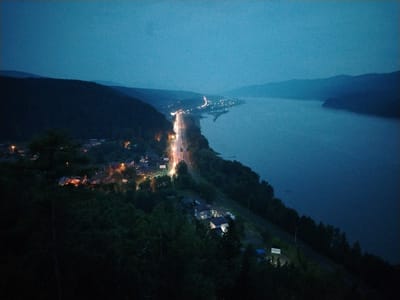 The image is a nighttime aerial view of a settlement along a body of water and a road. The scene is predominantly dark blue and black, with various lights from the settlement and the road illuminating parts of the landscape. In the foreground, dense dark green trees and foliage dominate the lower portion of the image, with some lights from houses peeking through. A dirt road or path is visible, with a few small structures and some parked vehicles. Further into the image, a well-lit road with traffic lights and car headlights stretches along the shoreline, bordered by a line of houses and buildings with illuminated windows. The body of water, likely a lake or river, takes up the right side of the image, appearing calm and reflecting the dim light. In the background, dark, silhouetted hills or mountains recede into the twilight sky. A white sign with black lettering, possibly Russian text, is visible on the lower right side of the image, near a small blue tarped structure. Based on the provided context, this scene is located in the city of Слизнево, Russia.