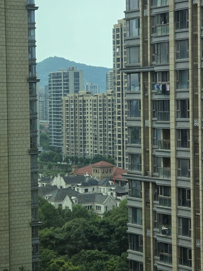The image shows a view of a residential area in Xiaoshan District, China, with several tall apartment buildings and some lower-rise housing. In the foreground on the left is a close-up of a light-colored brick apartment building with many windows. To the right and further back are two more modern apartment buildings with glass balconies. Laundry is visible hanging from a balcony on the rightmost building. In the middle ground, there are clusters of white houses with dark roofs, and beyond them, more apartment buildings. Lush green trees are interspersed throughout the scene, and a hazy, tree-covered mountain rises in the background under a pale sky.