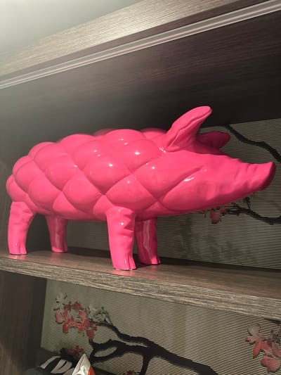 The image depicts a bright pink, stylized sculpture of a pig, possibly made of ceramic or plastic. The pig is in a standing position. Its body has a quilted or faceted texture. The sculpture is located on a shelf within a built-in cabinet or shelving unit. The cabinet appears to have dark wood or laminate doors and light-colored trim. The interior wall of the cabinet is covered in a decorative wallpaper featuring a striped pattern with floral elements. The image's focus is on the sculpture, with its surroundings providing context. There is no information given in the image to indicate the location of this scene within Genk, Belgium.