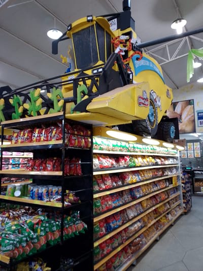 The image shows the interior of a supermarket in São Gabriel Do Oeste, Brazil, featuring a large, yellow, combine harvester decoration suspended from the ceiling. The combine is adorned with brand logos, including "ElmaChips" and "Supermercado São Gabriel." Below the combine, shelves are filled with various packaged snacks and food items, primarily chips and cookies. The shelves are constructed with black metal frames and light-colored wooden planks, illuminated by overhead lighting. In the background, there are more shelves stocked with products, a digital display showing prices, and a blurred image of baked goods on a wall. The floor is tiled with gray hexagonal tiles. There are no people clearly visible in the image.