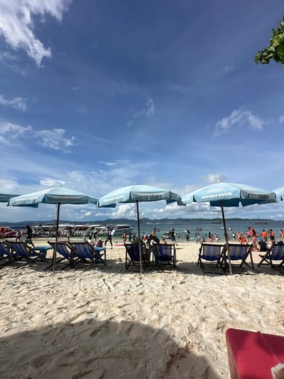 The image depicts a crowded beach scene on a sunny day in Phru Nai, Thailand. In the foreground, several empty lounge chairs with blue and striped cushions are set up on the white sand, arranged under light blue umbrellas. The umbrellas display the "JETSKI CLUB" logo. Beyond the chairs, a shallow area of the ocean is filled with people, some wading and others engaged in water activities. A fleet of speedboats and jet skis are docked or moving in the water near the shore. In the distance, a line of green, hilly islands forms the horizon under a bright blue sky with scattered white clouds. Numerous people are visible on the beach and in the water, some wearing life vests. The overall impression is a bustling tourist destination popular for water sports.