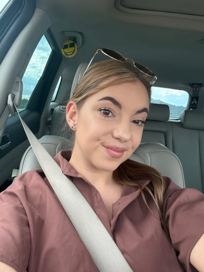 A young woman with brown hair pulled back and sunglasses perched on top of her head is taking a selfie inside a car. She is wearing a maroon collared shirt and a gray seatbelt. The interior of the car is visible, including the seats and the sky through the windows. A smiley face air freshener with sunglasses is hanging from the rearview mirror. The scene appears to be in Aldomirovtsi, Bulgaria.