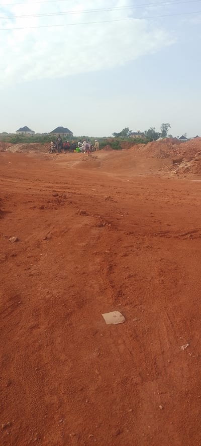 The image depicts an outdoor scene in Rayfield, Nigeria, characterized by a large area of reddish-brown earth, likely a construction or excavation site. In the mid-ground, a group of approximately ten individuals is gathered, seated and standing amidst piles of dirt. They appear to be taking a break, with some holding containers. In the background, several modern-looking houses with pitched roofs are visible, situated on a slightly elevated and vegetated area. The sky is partly cloudy and bright. The foreground is dominated by the textured, uneven earth surface.