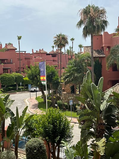 This is a vertical, eye-level, medium shot of a street lined with buildings and lush vegetation, captured on a clear, sunny day. The buildings, painted a distinctive terracotta hue, feature tiled roofs and appear to be apartments or resort accommodations. Palm trees and large-leafed tropical plants, such as banana trees, are abundant, creating a vibrant, almost jungle-like atmosphere around the buildings and along the street.
A paved road curves through the scene, bordered by green lawns and more trees. A pedestrian crossing sign is visible on the side of the road. In the mid-ground, a flagpole displays a flag with several colored stripes, including red, yellow, and green, alongside a blue section with what appear to be stars and a symbol. The flag is partially obscured by tree branches. The time of day appears to be midday, with bright sunlight casting distinct shadows, suggesting warm weather.
The scene is devoid of any visible people, focusing instead on the architecture and the natural elements. The overall impression is one of a serene, well-maintained resort or residential area in a warm climate, likely Spain, given the context provided. The clear sky and bright lighting evoke a sense of calm and pleasant weather. No discernible emotions are conveyed, as the image is purely descriptive of the setting. There is text on the flag, but it is illegible in this resolution.