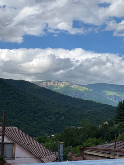 The image depicts a mountainous landscape with a village in the foreground, likely located in Dilijan, Armenia. A dense, dark green forest covers the lower slopes of a large mountain that dominates the mid-ground. The upper part of the mountain range features jagged, rocky peaks with patches of green vegetation, illuminated by sunlight filtering through dramatic, grey and white clouds. In the foreground, the weathered, rust-colored metal roofs of several houses are visible. One house on the left has a pinkish-beige exterior wall and a large window covered with a translucent curtain. A rough wooden pole stands to the left of this house. The sky above is a vibrant blue, with large, billowing clouds casting shadows on the mountains. The overall scene suggests a natural, somewhat rustic environment with signs of human habitation.