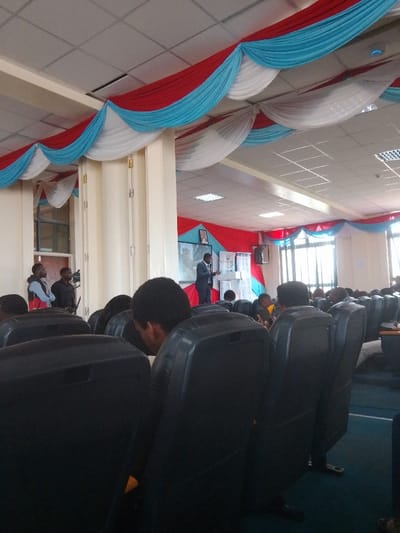 The image depicts an indoor event, likely a presentation or conference, taking place in Karingani ward, Kenya. The perspective is from the rear of an audience, looking towards the front of a spacious hall or auditorium.
**Environment:** The room features a white grid ceiling with numerous recessed lighting panels. Extensive decorative fabric drapes, in alternating red, light blue, and white, are festooned across the ceiling and along the upper walls. A tall, light-colored pillar is prominent on the left side of the room. The floor is covered with a blue carpet. Large windows are visible on the far right wall, admitting natural light. The front wall is partially adorned with similar red and blue fabric.
**People:** Approximately twenty individuals are visible, primarily from behind, seated in rows of dark, possibly black, high-backed padded chairs, facing the front. In the central foreground, the back of a person's head with dark, curly hair is prominent. At the front of the room, a male speaker dressed in a dark suit stands behind a white podium. To the left of the speaker, two additional male individuals are standing; one is operating what appears to be a video camera on a tripod, while the other stands nearby.
**Objects:** The white podium is centrally positioned at the front. Behind the speaker and podium, a large screen or display is visible, featuring an image, possibly a portrait or graphic. Wall-mounted speakers or monitors are visible on the front walls. The dark chairs are arranged in orderly rows.