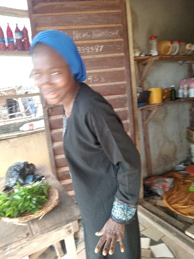 The image shows a young woman wearing a black dress and a blue headscarf. She is standing next to a wooden table with green leafy vegetables in a basket. The table is situated near a doorway, and the background shows a wooden shelf with various items on it. The woman's right hand is extended towards the camera. The location is a shop in Ota, Nigeria, with coordinates 6.691 latitude and 3.177 longitude.