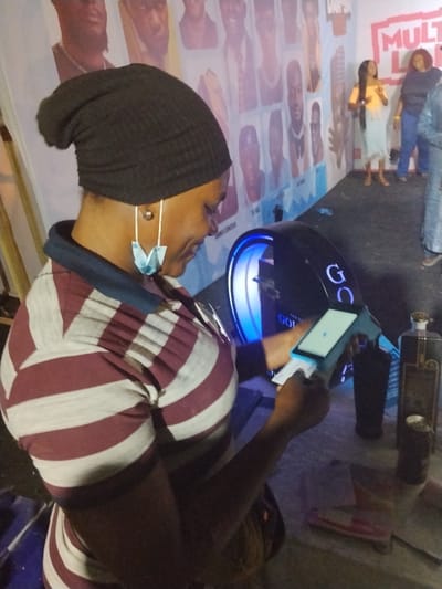 An indoor scene in Fagge D2, Nigeria, depicts a woman in profile view from the chest up, facing right and looking down. She wears a dark grey beanie and a maroon and white horizontal striped short-sleeved top with a dark collar. A light blue surgical mask hangs from her right ear, along with a white earbud. Her hands are positioned to hold a smartphone horizontally, and she appears to be interacting with its screen. A small white paper or card is visible in her left hand.
On a surface in front of her is a black, curved device with a blue illuminated interior and white text, "GOD," visible on its side. To the right of this device, two dark-colored bottles, one with a prominent stopper, and a silver-colored can are present on the same surface.
In the background, a light-colored wall is decorated with multiple framed portraits of men and text. Prominent red and white stylized text, "MULTILIVE," is visible on the upper right wall. Three individuals stand in the background on the right: one woman with long dark hair wearing a yellow off-the-shoulder top and a light skirt, and another woman in a dark blue top and jeans. A third person is partially obscured on the far right. The floor is dark.