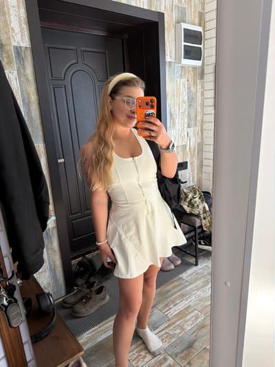 A woman is taking a selfie in front of a mirror. She is wearing a white dress and glasses, with a headband on her head. She is holding an orange phone in her hand.
The location is suggested to be in Большая Шильна, Russia.
The background features a wooden door, wood-look tiled walls, and a glimpse of the floor. There are shoes, bags and other items visible near the bottom and top right of the frame.