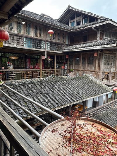 An elevated view captures a multi-story traditional wooden building complex in Gongtan, China, under an overcast sky. In the immediate foreground, a round, woven bamboo tray filled with dried red chili peppers and a branch bearing small red berries rests on a wooden railing. Three parallel, horizontal wooden poles extend across the lower-mid frame, situated above a dark grey tiled roof.

The architectural complex features multiple structures constructed primarily from dark timber, characterized by intricate wooden lattice windows, carved railings, and numerous dark grey traditional tiled roofs. Red lanterns are suspended from eaves at various points.

On an open-air wooden balcony of a central building, an adult, dressed in dark clothing, holds a child wrapped in a patterned blanket. Another individual is seated at a table on the same balcony. Multiple tables and chairs are visible arrangements. Red celebratory decorations, including one prominently displaying "2026" and others resembling traditional couplets, adorn pillars and walls on this balcony level.