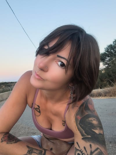 The image captures a woman in a casual outdoor setting during the golden hour, likely sunset or sunrise, given the soft, warm light and the gradient sky from light blue to pale orange at the horizon. She is positioned close to the camera, looking directly into the lens with a slight smile and a confident expression.
The woman has short, dark brown hair with bangs partially covering her forehead and her left eye. She has dark eyes, light pink lips, and a stud piercing in her right nostril and a ring piercing in her left. She is wearing a light purple bralette or bikini top, revealing several tattoos. A prominent abstract facial design is visible on her chest near her cleavage. Her left arm features a large, detailed tattoo that appears to be a hooded or veiled figure. Other tattoos are visible on her right arm and legs, though less distinct. She wears chain earrings with small charms on her left ear. A brown fanny pack or small bag is around her waist, and a glimpse of light-colored shorts or underwear is visible.
The background shows an open sky. A thin, dark cable or wire runs diagonally across the upper left portion of the sky. In the mid-ground, a dry, vegetated landscape with scattered bushes and trees stretches into the distance, consistent with a Mediterranean climate. The foreground suggests she is on or beside an asphalt or dirt road. No visible text is present in the image. The scene conveys a relaxed and natural moment.