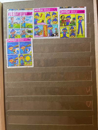 This is a photograph of a page from a sticker album, likely designed for children. The page is partially filled with colorful comic strip stickers. The album itself appears to be bound and is placed on a flat surface, possibly a table or shelf.
The visible stickers depict cartoon characters, primarily a boy with a blue hat and jacket and a pink shirt, and an older man with glasses and a green shirt. The illustrations are in a simple, child-friendly comic style.
One set of stickers, labeled "174" and "pembo çigniyor ve düșünüyor" (pembo is chewing and thinking), shows the boy in various poses, including one where he appears to be jumping and another where he is interacting with an older man in a workshop setting with a sewing machine. Another sticker set, labeled "170" and "pembo çigniyor ve düșünüyor", also features the boy, one panel showing him with a question mark above his head and a mouse with a briefcase. Below these are stickers labeled "169" and "pembo çigniyor ve düșünuyor", depicting the boy and another character in an outdoor, grassy setting. Finally, a sticker labeled "180" and partially obscured shows the boy looking through a telescope. One panel within this set has the word "AYDEDE" written on it. The stickers are arranged in a grid-like fashion.
The background of the album page is a dark, textured material with vertical indentations, suggesting a structured way to hold the stickers. The lighting is neutral, indicating an indoor setting, possibly during the daytime. The overall impression is of a collection of playful, cartoon-style stickers meant to be collected and displayed.