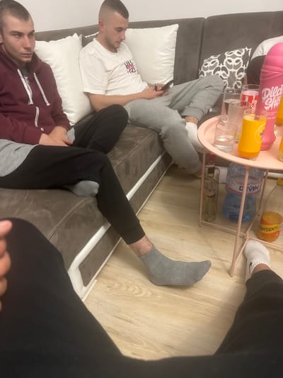 Three men are relaxing indoors. Two men are reclining on a couch, one wearing a white t-shirt and grey sweatpants, the other wearing a burgundy hoodie and grey sweatpants. The third man, partially visible, is wearing black pants. A side table with various beverages sits in front of the couch. The location is Graf Ignatievo, Bulgaria.