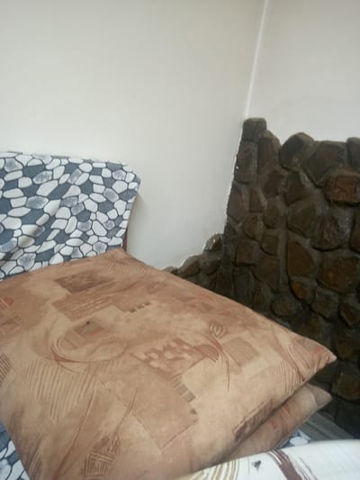 This media file shows a close-up view of two pillows in a room. The pillows are stacked, with the larger one in the foreground. The larger pillow has a faded brown fabric with a geometric pattern in shades of brown and cream. The smaller pillow underneath is mostly obscured but appears to have a similar pattern. Behind the pillows, to the left, is another pillow or cushion with a pattern of gray and white irregular shapes resembling pebbles. To the right, a wall made of rough, dark stones is visible. The background wall is a plain, light color, possibly white or cream, and the corner where the stone wall meets this plain wall is visible. The lighting suggests an indoor setting, with no specific time of day or weather conditions evident. There is no visible text in the image. The scene appears to be a domestic setting, possibly a living room or bedroom, in Lomets, Bulgaria.