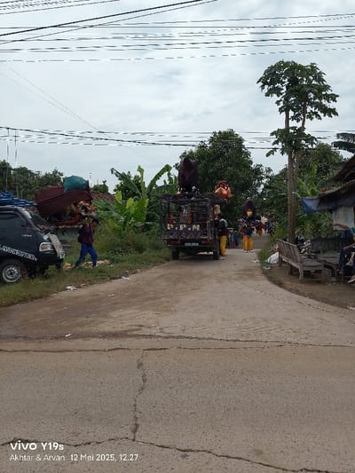 The image shows a paved road in Kecamatan Anjatan, Indonesia (latitude: -6.418, longitude: 107.968). There are several people standing around a truck that is loaded with various objects. Two people are walking on the road. The truck has a black canopy and is parked on the right side of the road. The road is cracked and there is a lot of debris on it. The environment is rural, with lush vegetation and a house in the background. There are electrical wires and a cloudy sky overhead. The scene appears to be a typical street in a small Indonesian town. The image is taken from a smartphone.