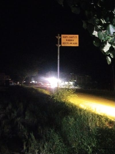 The image captures a nighttime scene in Meukokuthang, Indonesia. A rectangular yellow sign, mounted on a dark pole in the upper-middle left, displays three lines of black text: "HATI-HATI !!!", "RAWAN", and "KECELAKAAN" (Indonesian for "BE CAREFUL !!! ACCIDENT PRONE AREA"). The foreground is covered with dense, dark green vegetation and grass. A strong yellow-white light illuminates a portion of the ground and vegetation along the right side, extending from the mid-ground to the lower right. In the mid-ground, a road is visible with multiple vehicles whose bright white headlights create significant glare. Dark silhouettes of trees and indistinct buildings or structures are present behind the vehicles on the left. The sky is uniformly black. Dark green foliage, likely tree branches and leaves, partially frames the upper right corner of the image. No individuals are discernible.