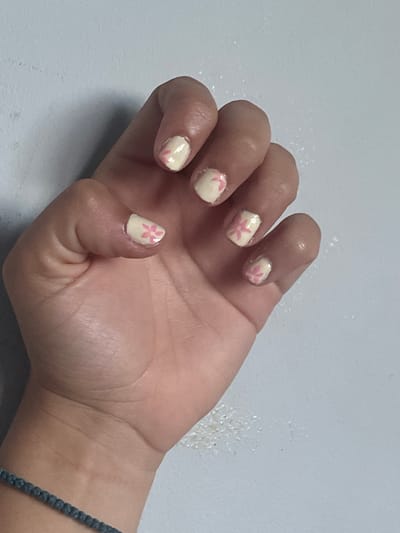A human hand, likely female, is prominently featured, resting with its palm facing upwards on a light-colored, plain surface. The primary focus is on the fingernails, all of which are painted with a pale cream or light yellow base coat. Each visible nail (thumb, index, middle, ring, and pinky) is decorated with small, simple pink floral designs, resembling cherry blossoms or similar delicate flowers.
Tiny, sparkling specks, possibly glitter from nail polish or residual particles, are visible on the skin around the cuticles and on the light grey or off-white surface beneath the hand. The skin of the hand appears smooth, and the natural lines of the palm are visible. On the wrist, to the right side of the image, a dark-colored, possibly braided or beaded bracelet is worn. The setting appears to be indoors, under soft, even lighting, without any specific time of day discernible. The overall activity depicted is simply the presentation of the hand and its adorned nails. No text is visible in the image. This scene is located in Maidstone, United Kingdom.