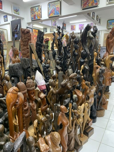 An indoor art gallery or shop in Ikate, Nigeria, is densely filled with a vast collection of wooden sculptures and carvings. The foreground and midground are dominated by hundreds of figures crafted from various types and shades of wood, ranging from light brown to dark, almost black, tones. Many sculptures depict stylized human forms, primarily female figures, featuring elongated necks, intricate hairstyles, and full body or torso representations. Abstract and more realistic forms are also present. These sculptures are tightly packed, arranged on the white tiled floor and low display platforms. The white walls of the establishment are adorned with numerous framed paintings of various sizes and styles, including abstract, landscape, and figurative works, characterized by vibrant colors. Overhead fluorescent lighting illuminates the entire scene.