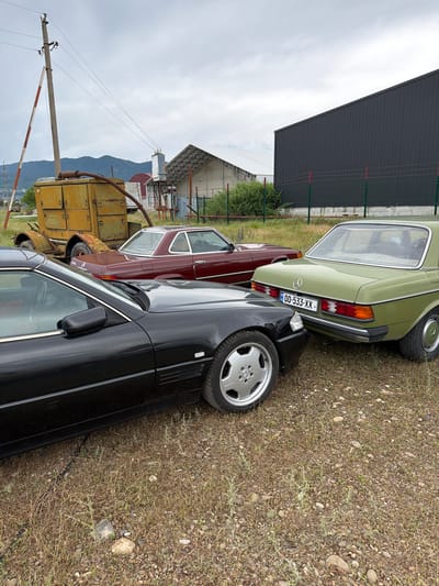 In Natakhtari, Georgia, three vintage Mercedes-Benz cars are parked on a gravel and dry grass lot. The cars are a black coupe in the foreground, a maroon coupe in the middle, and a light green sedan in the background. Behind the cars, a large dark grey building with a corrugated metal facade stands, fenced off by a green metal fence with red posts. To the left, a rustic yellow generator and a utility pole with wires are visible, with a mountainous landscape and a cluster of industrial buildings in the distance under a cloudy sky.