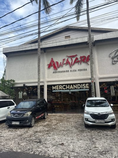 This outdoor image depicts a commercial building in Kampung Toyapakeh, Indonesia, hosting an "Awatar" merchandise shop. The building's facade features a large red "Awatar" sign above the word "Merchandise," with palm tree trunks framing the building's front. Two cars are parked in front of the shop: a white car with the license plate "DK 1942 FCD" and a black car with the license plate "DK.1579. AO." Power lines and palm fronds obscure part of the sky above the building. The ground appears to be paved with stone. The image is oriented vertically.
