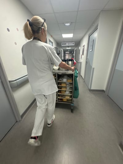 The image depicts a corridor, likely within a healthcare facility in Tourcoing, France. A person wearing white uniform (possibly a nurse or healthcare worker) is standing beside a medical cart filled with supplies. The corridor is painted in neutral tones with visible signage and lighting fixtures on the ceiling. The floor has a patterned surface.