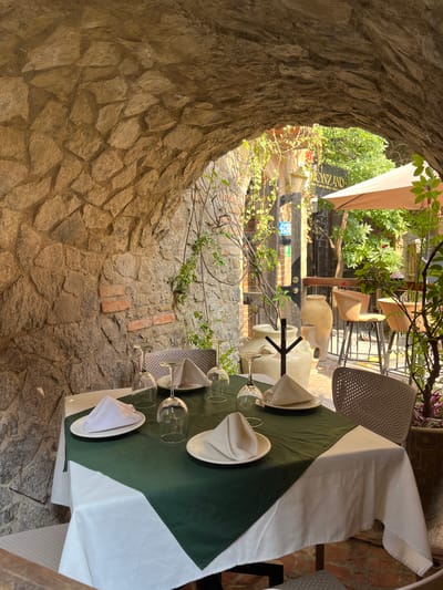 The image shows a set table at an outdoor restaurant in Domingo Arenas, Mexico. The table is covered with a green and white tablecloth and set with plates, napkins, and wine glasses. The restaurant is located in a stone structure with an arched opening that looks out onto a courtyard. The courtyard contains additional tables, chairs, and plants. A sign for the restaurant is visible in the background. There are no people visible in the image.