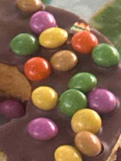 The image displays a close-up, overhead view of a chocolate cake decorated with colorful candies. The candies are arranged in a somewhat haphazard pattern across the dark chocolate surface, which appears to be slightly textured. Visible candy colors include purple, green, yellow, orange, and brown. Some candies are round, while others are slightly oval. The overall impression is that of a confectionery item, possibly a dessert. The image does not contain any contextual information or visual elements that would place it in the city of Heniivka, Ukraine, or any other specific geographical location. There are no people, actions, or interactions depicted in the scene.