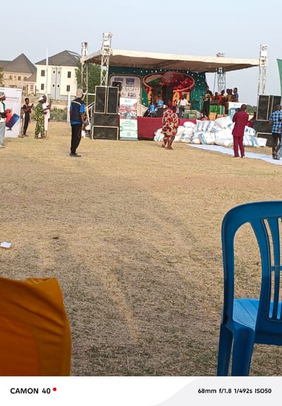 Here's a detailed description of the scene:
**Setting and Environment:**
The image captures an outdoor event, likely a festival or a public gathering, taking place in Fagge D2, Nigeria. The foreground consists of a grassy field, slightly barren with patches of dry vegetation. To the right is a blue plastic chair.
**Subjects and Objects:**
* **Stage:** In the mid-ground is a temporary stage structure, equipped with speakers stacked on either side. A banner or backdrop adorns the back of the stage, featuring vibrant colors and what appears to be cartoonish or artistic representations. A band is playing on the stage.
* **People:** Several individuals are scattered around the stage. They are diverse in attire, suggesting a mix of casual attendees, performers, and possibly organizers. Some are dressed in traditional Nigerian clothing.
* **White Bags:** Piles of white bags (likely filled with goods) are arranged in front of the stage, serving as a barrier or perhaps part of the event's logistics.
* **Banner:** To the left, there is a banner for the Tecno Camon 20 series.
* **Buildings:** In the background, residential or commercial buildings are visible.
**Actions and Interactions:**
* **Band Performance:** A band is actively performing on stage, suggesting an ongoing entertainment segment.
* **Interaction:** People are standing around the stage, presumably watching the performance.
**Overall Impression:**
The image conveys a sense of community and cultural celebration. The event has drawn a crowd, and the atmosphere seems festive and engaging. The stage structure and musical performance indicate a planned event. The barren field setting suggests an open-air gathering in a less urbanized area.