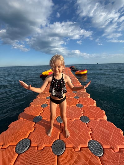 A young girl with blonde hair and a black and white polka dot bikini stands on an orange floating dock. She is facing the camera with her arms outstretched, palms up. The dock is made of interlocking hexagonal modules and extends into dark blue water. In the background, a red and yellow inflatable object floats on the water. The sky above is blue with fluffy white clouds. The sun is shining, indicating it is daytime. The scene appears to be a recreational area on the water, possibly a lake or the sea, near Yalta, Ukraine. The girl looks happy and excited. There is no visible text in the image.