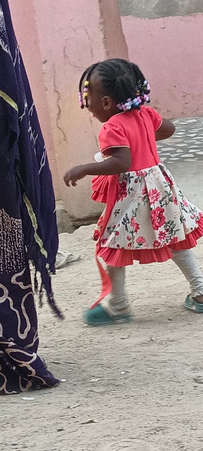 This vertical full shot depicts an outdoor scene in Rangaza, Nigeria, featuring a young child in motion, viewed from behind and slightly to the right, with a partially visible adult on the left. The child, likely a girl, wears a bright red, short-sleeved top and a patterned skirt that is predominantly white or cream with bold red and green floral designs. Underneath the skirt, red pleated ruffles are visible. She has light grey leggings and turquoise or teal slippers on her feet, with the left foot appearing blurred, suggesting movement. Her dark hair is styled in numerous braids, adorned with colorful beads in yellow, pink, purple, and light blue.
To the left of the frame, the lower body and midsection of an adult are visible, clad in a dark blue fabric with intricate white or light-colored swirling patterns and fringes, possibly a traditional garment. The background consists of a plain, light pinkish-brown wall, suggesting a building, while the ground is sandy or dusty. The natural lighting indicates it is daytime, likely morning or late afternoon. The overall setting and attire are consistent with a local environment in Rangaza, Nigeria. No specific emotions are discernible as faces are not clearly visible, and there is no visible text in the image.