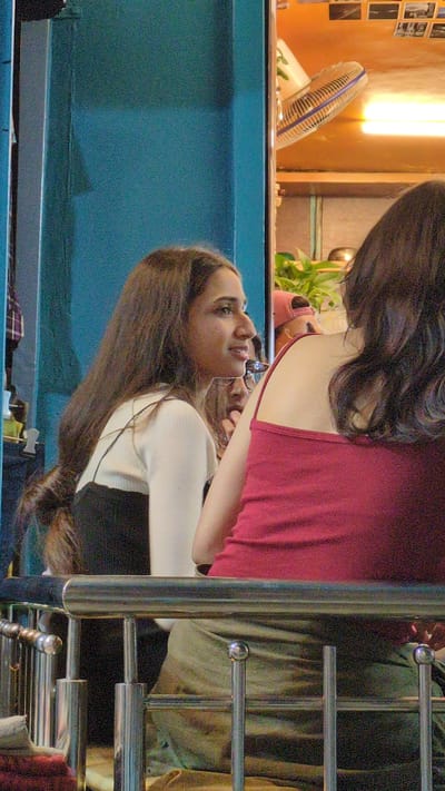 Captured indoors in what appears to be a living or dining area in Dharamkot, India, the photograph portrays two females. One female, dressed in a red sleeveless dress, stands partially visible in the upper right quadrant, her dark hair cascading down. The second female, with long brown hair, is reflected in a blue-surfaced table or countertop, wearing a white top with a black lower garment. She appears to be conversing with the other female. The reflection shows her wearing glasses. In the background, there is a small electric fan, some plants, and various indistinct objects, suggesting a lived-in space. A silver-colored metal railing adds a structural element to the scene. The image orientation is rotated 90 degrees counterclockwise.
