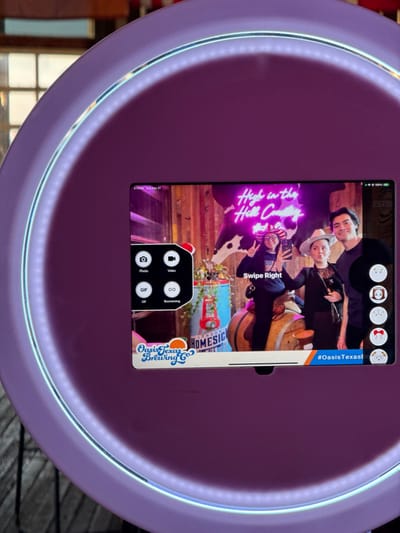 A purple photo booth captures a digital display showing four people posing with a neon sign reading "High in the Hill Country." One person holds a phone, presumably taking the picture. A "Swipe Right" prompt is visible. The bottom of the display advertises "Oasis Texas Brewing Co" and includes a London Homesic reference. The phone interface offers photo, video, GIF, and Boomerang options. Hashtags associated with the location, such as "#OasisTexasi," are also displayed. The image is taken from the photo booth itself, showcasing its built-in screen and lighting.