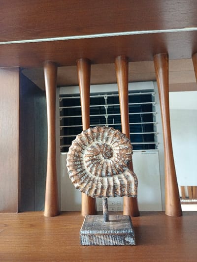A decorative fossil sculpture sits on a wooden surface. The sculpture is a spiral shape with ridges, mounted on a small square base. Behind the sculpture, a wooden structure with evenly spaced, rounded support beams frames a white air conditioning unit with a dark grill. The background is a slightly blurred, light-filled interior. The color palette is dominated by warm tones of brown and white.