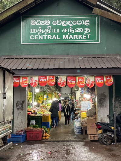 The image shows the entrance to the Central Market in Nuwara Eliya, Sri Lanka. A green sign displays the market's name in multiple languages. Above the entrance, red and white "Viva" advertising banners are affixed. Inside, a bustling market scene is visible with people shopping among displays of fruits and vegetables stacked in crates. A motorbike is parked outside the market. The ground appears wet, possibly from recent rain.