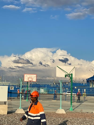 The image depicts an outdoor scene with a clear blue sky and dramatic cloud formations. In the foreground, a man wearing an orange hard hat and a high-visibility jacket walks on a gravel path. Behind him, a chain-link fence topped with barbed wire separates the foreground from a facility. Two basketball hoops are visible, one with a red rim and backboard, and another with a white backboard and green supports. Beyond the fence, several buildings painted blue are visible, along with other individuals, some wearing work attire, and a white car. The overall environment suggests an industrial or recreational area, possibly within or near the city of Koskolovo, Russia.