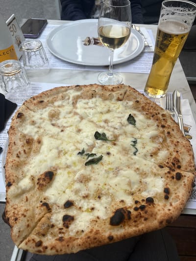 This media file presents an overhead view of a table set for a meal in Turin, Italy. The central subject is a large, round white pizza (pizza bianca), generously topped with melted white cheese, likely mozzarella or provola, with a few green basil leaves scattered across its surface. The pizza crust is thick, airy, and features characteristic dark charring, indicative of a high-temperature oven, typical of Neapolitan-style pizza. A light drizzle of olive oil is also visible on the cheese.
Accompanying the pizza are two beverages: a tall glass of light amber beer with a foamy head, and a stemmed glass containing white wine. The table is laid with a white plate bearing a partial logo, a set of silverware (fork and knife), and several small, empty drinking glasses. Papers with visible text, presumably menus or receipts, are spread across the white marble-patterned tabletop. Partially visible are a mobile phone and dark fabric, suggesting a person is seated at the table.
Visible Italian text on the papers includes "Pizzeria," "dolci" (sweets), "Casa" (house), "Provola" (a type of cheese), "Agerola" (a town known for cheese), "Pecorino" (another type of cheese), and "DOP cheese," along with a price of "3€." These details reinforce the Italian culinary context. The lighting suggests the photograph was taken during the daytime, capturing a casual dining experience.