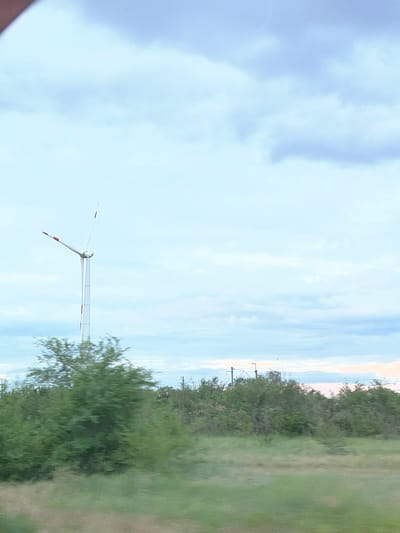 The image captures a landscape scene with a wind turbine prominently featured on the left side. The wind turbine is white with red accents on its blades and a tall, slender tower. It is surrounded by lush green trees and vegetation. The sky above is filled with a mix of clouds, appearing soft and diffused. In the background, the sky transitions to lighter hues, with hints of pink and orange suggesting either sunrise or sunset. The overall impression is one of a rural or semi-rural environment, possibly on the outskirts of a town or city. Based on the provided information, this scene is located in the city of Zimovniki, Russia.