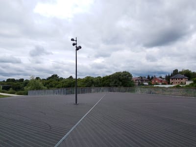 The image depicts an outdoor scene on a cloudy day, likely in the afternoon. In the foreground, a dark, textured wooden platform or deck stretches out, with a distinct diagonal line composed of a metal strip bisecting it. A tall, dark lamppost with multiple light fixtures stands on the left side of the platform, near a thin metal railing that extends horizontally across the middle ground.
Beyond the railing, lush green trees form a dense backdrop, with a weeping willow visible on the left. In the distance, nestled amongst the trees, are several red-tiled roof houses with dark brickwork, suggesting a residential area. The sky is overcast with dramatic, grey clouds, indicating a cool or potentially rainy weather.
The scene appears to be a public space, possibly a park or a viewpoint overlooking a natural area with some man-made structures in the background. There is no visible text in the image, and no people are present, contributing to a sense of quiet solitude. The overall mood is calm and somewhat somber due to the cloudy sky. The location is identified as Orekhovo-Zuyevo, Russia.