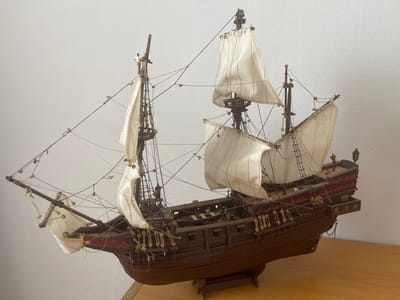 The image displays a highly detailed model of a wooden sailing ship, likely a historical replica, positioned on a wooden surface against a plain, off-white wall. The ship is brown with red decorative accents along its hull and upper decks. It features multiple masts with cream-colored sails that are unfurled and rigged with numerous thin lines, simulating the complex rigging of a real vessel. Several small, unpainted figures are visible on the ship's decks and rigging, suggesting miniature crew members. A small dinghy is depicted on the main deck, and other miniature accessories like cannons are present along the sides. The ship rests on a wooden stand. There is no discernible location in the city of Las Palmas, Spain, indicated in the image. The scene is static, showing the model ship at rest.