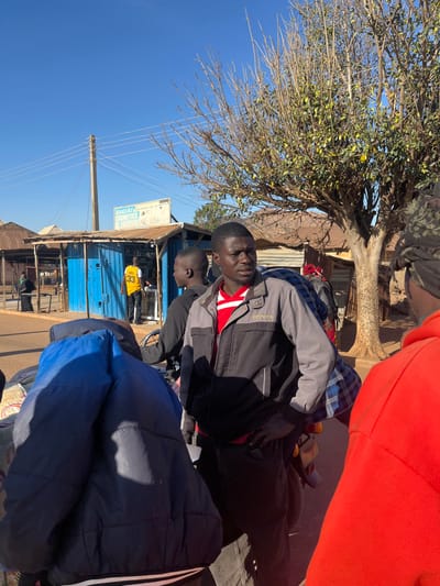 Several people stand outdoors with a tree overhead and a blue sky visible. A building with a sign that reads "BAKERIES, COSMETICS & FASHION" is visible. Two men stand close together, one wearing a gray and black jacket with a red shirt underneath. Others are partially visible, wearing an orange shirt and a blue jacket. The scene appears to be a street or marketplace.