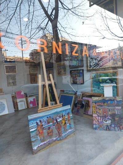 The media file captures a reflected view into what appears to be an art gallery or shop window on a street in Tiranë, Albania. The image is rotated 90 degrees clockwise, showing the reflections and interior contents vertically.
Reflected in the window, dominating the left side of the frame, are the intricate, bare branches of a large tree set against a bright, overcast sky. This suggests a winter or early spring day. Faint architectural details of a building facade are also visible in the reflection, hinting at an urban setting.
Inside the shop, numerous paintings are displayed. One prominent painting, positioned on a wooden easel, depicts several human figures swimming or floating in a vibrant blue body of water, possibly a beach scene, executed in a thick, impressionistic or expressionistic style. Another sizable painting, leaning against a wall or stacked, features a colorful townscape with distinct buildings and perhaps a harbor or body of water. Many other framed and unframed artworks of various sizes and subjects are visible throughout the interior, some stacked on the floor, others hung on walls. The floor inside appears to be a plain, light-colored surface.
A notable detail is the orange text, reflected and reversed, visible across the window. When de-reflected and oriented correctly for external viewing, it reads "OR NIZAZ & H". This is likely part of the business name for the establishment. The overall scene suggests a quiet daytime street view interacting with an indoor artistic display.