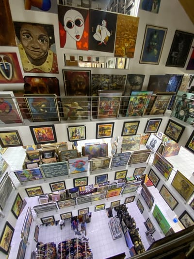 This image captures a high-angle, overhead view of a multi-level art gallery located in Ikate, Nigeria. The interior features white walls and white tiled flooring on the ground level. Numerous framed and unframed paintings densely cover the walls from the ground floor up to the visible ceiling, with additional artworks stacked on various shelves and racks across multiple intermediate levels. Metal railings delineate these upper floors. The displayed art encompasses diverse styles, including portraits, abstract figures, landscapes, and genre scenes, executed in a variety of vibrant colors. On the ground floor, three distinct individuals are visible. Two men, one in a light blue collared shirt and dark trousers, and another in a light-colored shirt and dark trousers, stand near a display of neatly folded, colorful textiles or rolled canvases arranged on low platforms. A third person in a red garment is present nearby. To the right, a multi-tiered display holds numerous dark-colored sculptures or artifacts. The entire space is brightly illuminated, indicating ample artificial lighting.