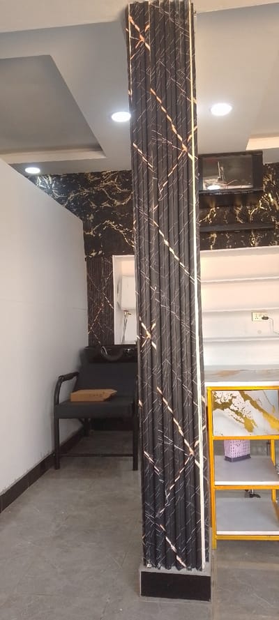The image displays the interior of a commercial establishment, specifically a salon or beauty parlor, located in Matathani ward, Kenya.

A prominent, dark-colored structural pillar with a vertically fluted or ribbed texture, adorned with irregular diagonal lines in a light gold or copper tone, extends from the grey tiled floor to the white ceiling. The floor tiles are large and exhibit subtle lighter veining. The white ceiling features a recessed panel design and contains multiple circular LED downlights.

To the left of the pillar, a black upholstered chair designed for hair washing, complete with armrests and an integrated dark-colored basin, is positioned against a wall. This wall is clad in dark paneling featuring a distinct gold-veined marble pattern. A rectangular mirror is partially visible on this patterned wall, situated behind the washbasin. A flattened, unsealed brown cardboard box rests on the seat of the hair-washing chair. The wall section immediately to the left of the washbasin area is plain white.

To the right of the central pillar, a multi-tiered shelving unit with a gold-colored frame and grey shelves is visible. The top shelf of this unit holds a rectangular item with a gold frame, displaying an abstract pattern of white and gold elements, alongside a light purple woven basket. Above this shelving, a dark-colored television or monitor is mounted on the gold-veined patterned wall; its screen shows an indistinct image, and visible wires extend above it. A white electrical outlet with two sockets is present on a white wall section to the right of the shelving unit.

No individuals are present, and no actions or interactions are depicted within the scene.